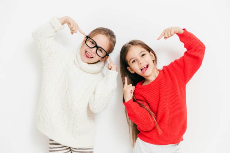 Joyful girls being in good mood, wear oversized sweater and spectacles, make cool sign, pose together against white background. Happy female children have fun. Childhood and happiness concept